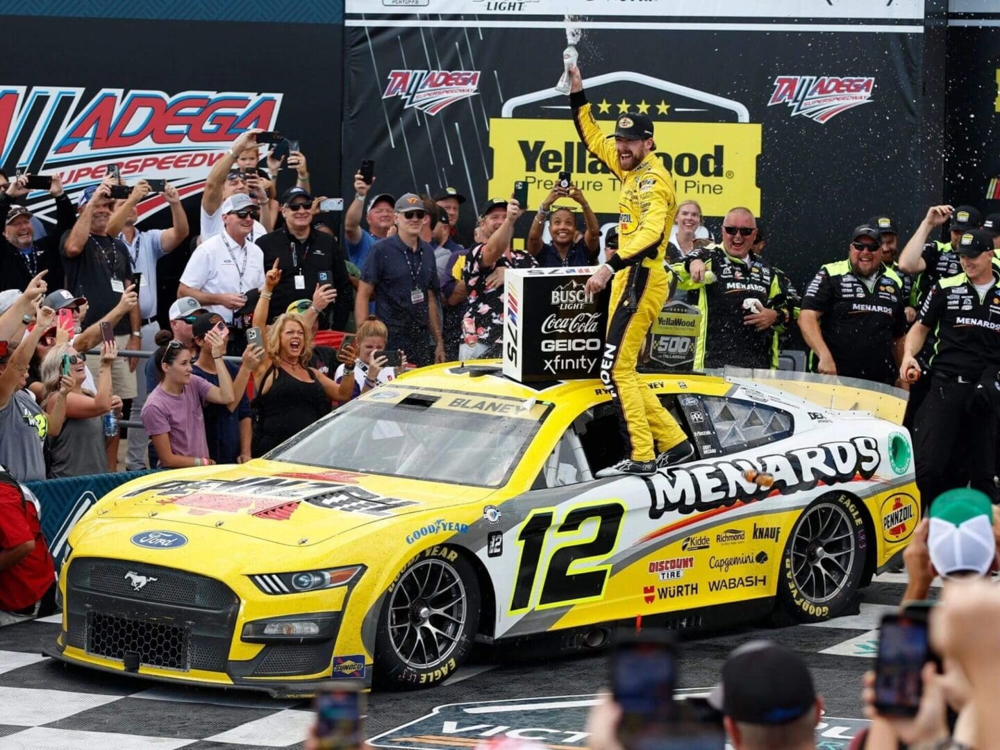 A man in yellow racing suit standing on the hood of a race car.